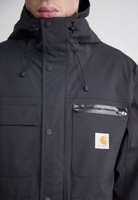 Zwarte waterdichte jas met een hoge kraag, twee voorzakken, een ritsvak op de borst en een logopatch. Voorzien van verstelbare koordjes.