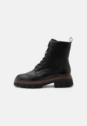Černá kožená kotníková šněrovací bota s prošíváním brogue, silnou podrážkou s výrazným vzorkem a poutkem na zadní straně, zobrazena ze strany na bílém pozadí.