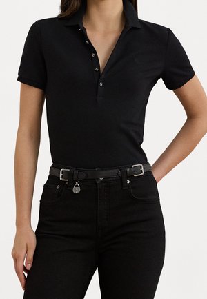 Vrouw draagt een zwart poloshirt met korte mouwen en knopen, en zwarte jeans met een zwarte riem met een kleine zilveren hanger.