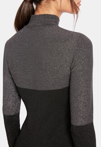 Femme portant une robe à manches longues, col haut et paillettes, avec une partie supérieure grise et une partie inférieure noire, vue de dos et en profil.