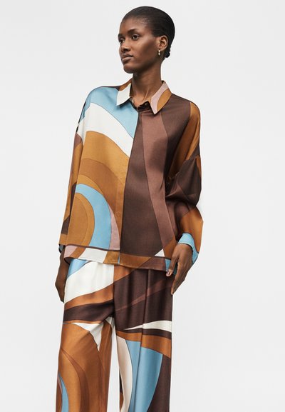 Nainen, jolla on löysät sopivat paita ja housut, joissa on abstrakteja ruskeita, sinisiä, valkoisia ja beige geometrisia kuvioita valkoisella taustalla.