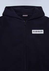 Felpa con zip di colore navy realizzata in un tessuto morbido. Presenta una tasca frontale e un'etichetta quadrata con logo sull'area sinistra del petto. Cappuccio foderato con una texture coordinata.