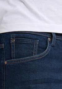Vaqueros de mezclilla azul oscuro con un bolsillo frontal, detalle de botón plateado y costuras contrastantes a lo largo de los bolsillos y la cinturilla.