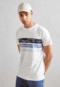 Weißes Baumwoll-T-Shirt mit horizontalem Design aus marineblauen und blauen Streifen, rundem Halsausschnitt und kurzen Ärmeln. Kombiniert mit einer hellbeigen Kappe.