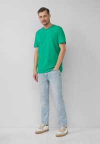 Zielona bawełniana koszulka z okrągłym dekoltem, zestawiona z jasnoniebieskimi dżinsami i białymi trampkami z beżowymi akcentami oraz sznurowadłami.