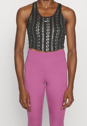 Personne en forme portant un débardeur court noir à motif doré et un legging taille haute mauve, debout devant un fond clair uni.