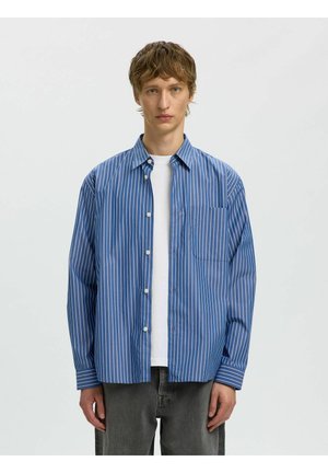 Jeune homme portant une chemise rayée bleue et blanche déboutonnée sur un t-shirt blanc et un pantalon gris foncé, debout devant un fond uni.