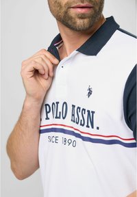 Hvid polo skjorte med marineblå krave, der har en rød og blå vandret stribe. Logoer og tekst i marineblå på fronten. Korte ærmer.