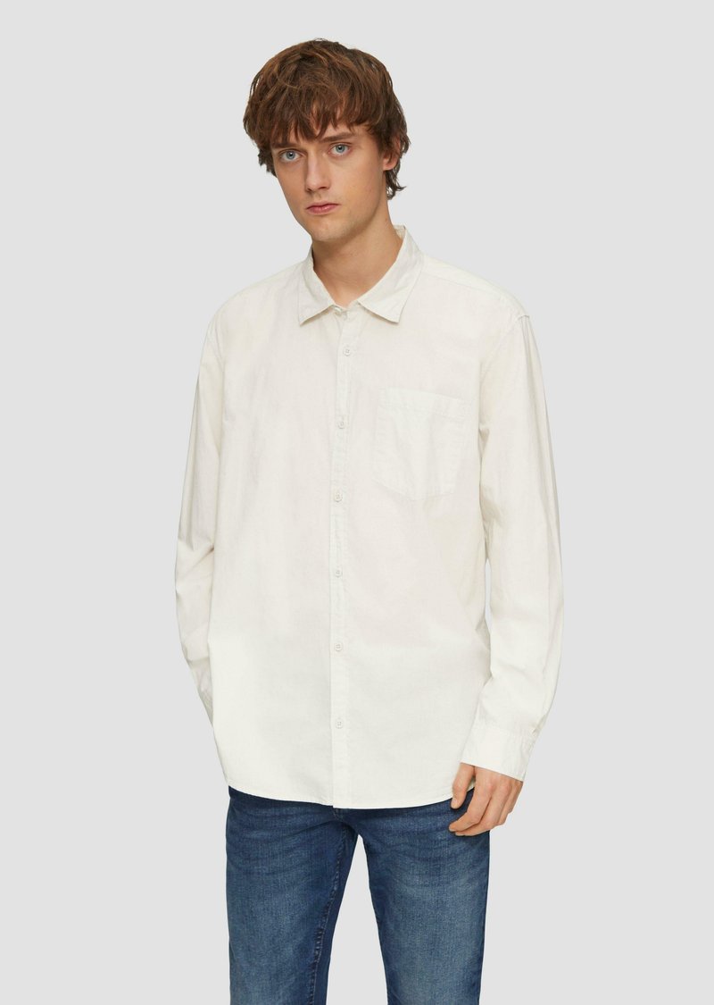 Chemise blanche à manches longues et boutons en coton avec une poche sur le côté gauche, portée sur un jean bleu, dotée d'un col classique et d'une coupe décontractée.
