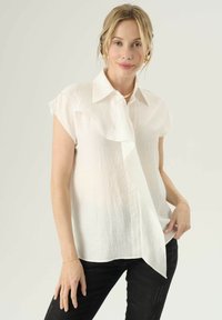 Blusa bianca a maniche corte con un davanti arricciato, design con bottoni e tessuto leggermente trasparente. Indossata con pantaloni neri.