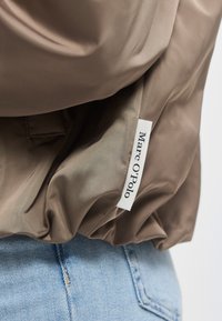 Brūns satīna žaketes piedurkne ar baltu Marc O’Polo etiķeti, apvienota ar gaisziliem džinsa džinsiem, kas redzami apakšā.