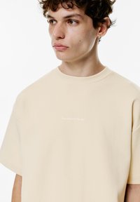 Kortærmet t-shirt med rund hals i lys beige bomuld, prydet med en kontrastfyldt teksttryk "Tættest på himlen" på brystet.