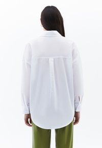 Camicia bianca a maniche lunghe con colletto classico, piega sul retro e orlo arrotondato; abbinata a pantaloni verdi. Il tessuto appare leggero e liscio.