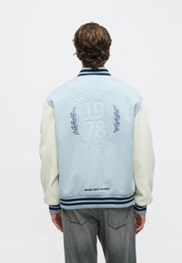 Άνδρας που φοράει ανοιχτό μπλε και κρεμ varsity μπουφάν με τα κείμενα "Carlo Colucci 1978" και "Broke Since Milano" στην πλάτη, στέκεται μπροστά σε λευκό φόντο.