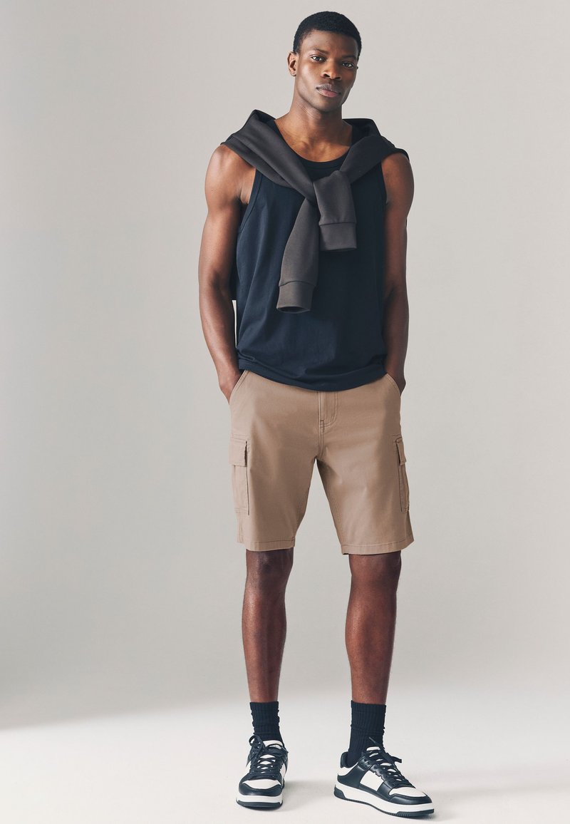 Débardeur noir avec un sweat-shirt marron drapé autour du cou, associé à un short cargo beige et des baskets noires à accents blancs.