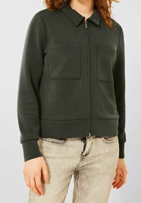 Giacca a zip verde scuro con colletto, due tasche frontali, polsini a costine e texture liscia. Abbinata a pantaloni in denim chiaro.