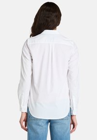 Camisa blanca de manga larga en algodón ligero, con yugo en la parte posterior, dobladillo redondeado y puños abotonados. Textura suave con mínimos detalles.