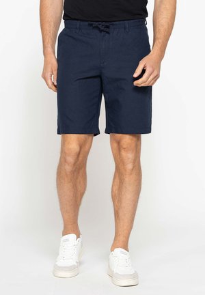 Uomo che indossa pantaloncini blu navy con coulisse e scarpe da ginnastica bianche, in piedi davanti a uno sfondo semplice.