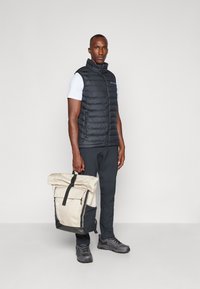 Sort puffer vest, hvid kortærmet skjorte, sorte bukser, grå sneakers og en beige og sort roll-top rygsæk med lynlås.