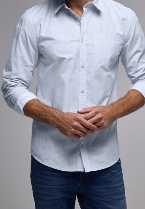 Mand iført lyseblå skjorte med knapper og opklebede ærmer og mørkeblå jeans, hænderne foldet ved taljen, neutral grå baggrund.