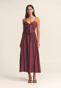 Bordowa sukienka w groszki z dopasowanym gorsetem, ozdobą w formie kokardki przy dekolcie oraz zwiewną, sięgającą do kostek spódnicą; cienkie ramiączka typu spaghetti.