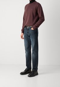 Sudadera burdeos con cuello redondo y pequeño logo, combinada con jeans azul oscuro y zapatos negros de cordones. Tejido suave, estilo ajustado.