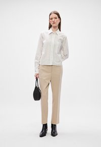 Camisa abotoada com riscas verticais em branco e cinza claro, combinada com calças bege ajustadas. Modelo segura uma pequena bolsa de mão preta.