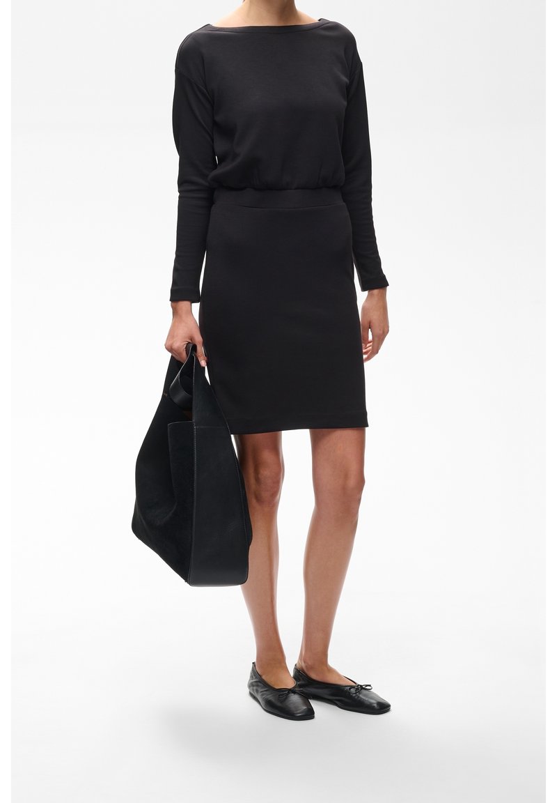 Femme portant une robe noire à manches longues et des ballerines noires, tenant un grand sac cabas noir sur un fond blanc.
