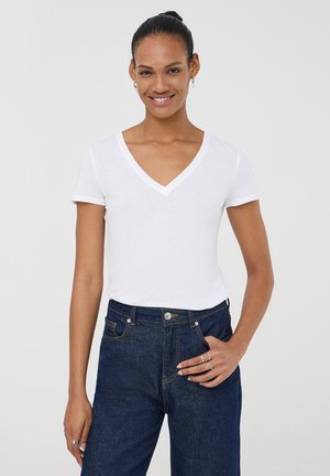 T-shirt bianco a maniche corte con scollatura a V in tessuto leggero, abbinato a jeans in denim blu a vita alta; il modello sta in posizione casual con le mani sui fianchi.