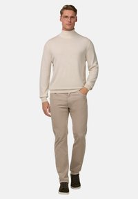 Krems krūšu izgriezuma džemperis, beigu krāsas chinos bikses, tumši zaļa sporta apavi ar baltām zoles. Vīrietis stāv taisni ar rokām kabatās, valkājot brilles.