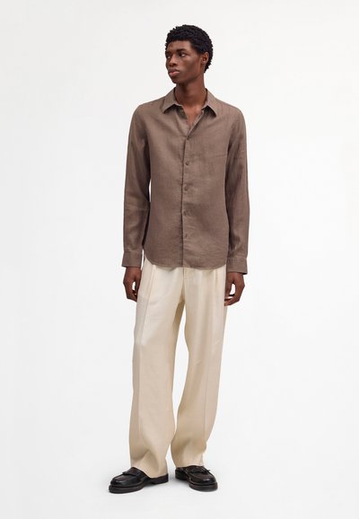 Homme portant une chemise marron à manches longues boutonnée, un pantalon crème à jambes larges et des mocassins noirs, debout devant un fond blanc uni.