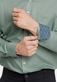 Camicia button-up verde chiaro e testurizzata con un polsino interno blu a fantasia. Presenta una tasca sul petto e bottoni bianchi. Sono visibili dei pantaloni neri.