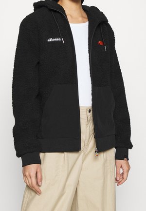 Person wearing a black zip-up fleece jacket with hood, "ellesse" logo, white top, and beige wide-legged pants, hands relaxed by sides.