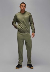 Ung man som står och bär en olivgrön Jordan-träningsdräkt med svarta och bruna sidostripes och svarta sneakers med gummisulor.