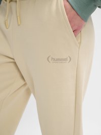Beige Hummel sportbroek met koordsluiting in de taille en een hand die in de rechtervoorzak rust tegen een witte achtergrond.