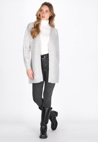 Ljusgrå stickad cardigan med öppen front, långa ärmar och ribbade kanter, kombinerad med åtsittande svarta jeans och knähöga svarta stövlar.