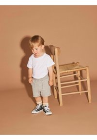 Niño con una camiseta blanca sencilla, pantalones cortos grises con cordón y zapatillas negras. Hay una silla de madera cerca sobre un fondo beige.