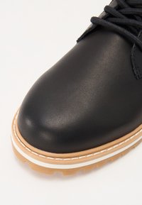 Black leather boot with a rounded toe, featuring a tan and white rubber sole and contrasting stitching along the edge.