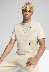 Beżowy t-shirt z krótkim rękawem, z kieszonką na zamek, czarnym logo Pumy na piersi, gładką tkaniną i luźnym krojem. Stylizowany z pasującymi spodniami.