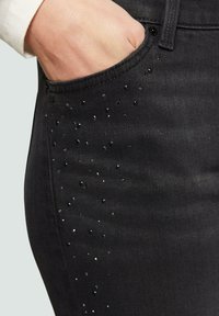 Jean en denim noir avec une coupe ajustée, ornés de clous noirs et argentés le long des poches. Taille mi-haute, couture classique.