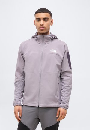 Νέος άνδρας που φορά ανοιχτό γκρι μπουφάν με φερμουάρ και κουκούλα και γκρι παντελόνι, στέκεται μπροστά σε απλό φωτεινό φόντο.