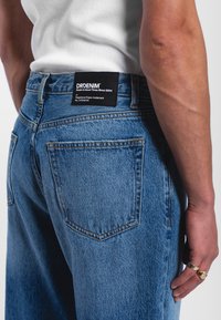 Light blue denim jeans with a relaxed fit, featuring fading, two back pockets, contrasting orange stitching, and a black branded label on the waistband.