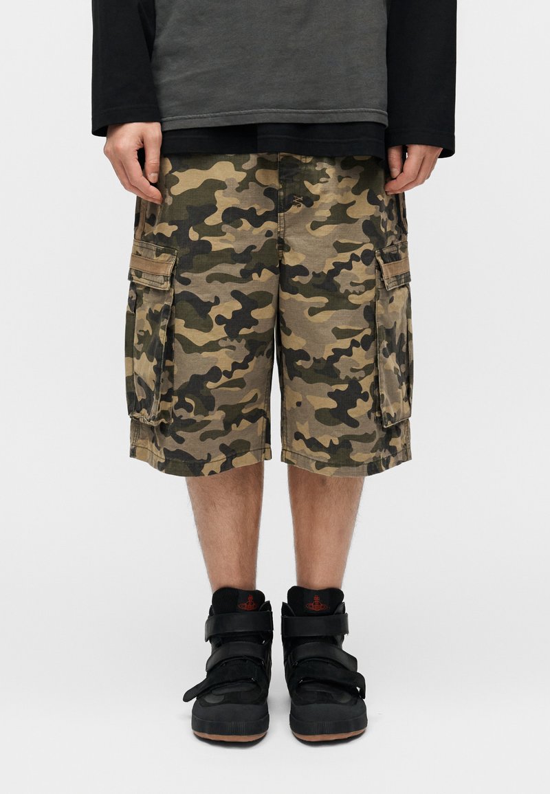 Cargo σορτς με καμουφλάζ μοτίβο, διαθέτουν πολλαπλές τσέπες, άνετη εφαρμογή και μήκος έως το γόνατο. Φορεμένα με μαύρα sneakers ψηλού τύπου.
