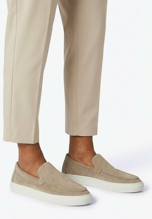 Beige Wildleder-Slipper mit weißen Sohlen, getragen zu hellbeigen, gekürzten Hosen, die nackte Knöchel zeigen.