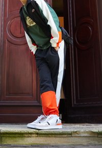 Träningsoverall med svart överdel och vita och oranga detaljer samt elastiska ärmar, ihop med vita sneakers med svarta och oranga inslag.