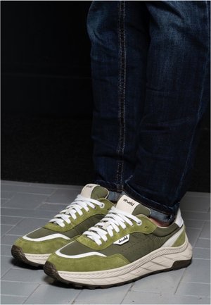 Personne portant des baskets vert olive et blanches avec des lacets blancs, associées à un jean bleu foncé, debout sur un sol carrelé gris clair.