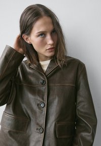 Brun læderjakke med knappefront, spids krave og to påsyede lommer. Glat tekstur med kontrastsyede detaljer.