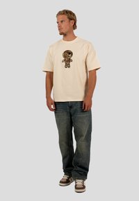 Beige Baumwoll-T-Shirt mit einem mehrfarbigen Cartoon-Charakter-Graphic, kombiniert mit lockeren blauen Denim-Jeans und braunen Sneakern.