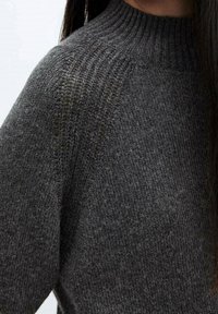 Donkergrijze gebreide trui met een hoge, geribbelde hals. De stof heeft een zachte textuur en een subtiel patroon, wat warmte en comfort benadrukt.