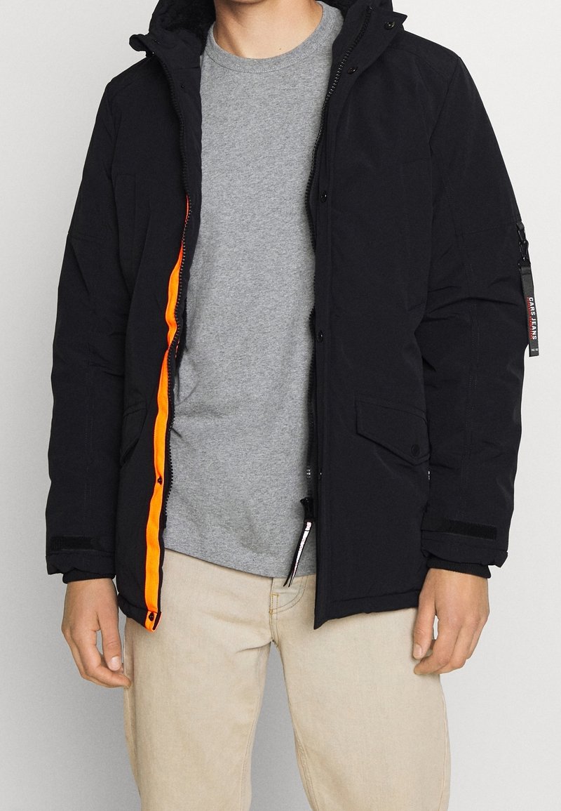 Svart isolerad jacka med en orange dragkedjedetalj, huva och kardborreknyten, bärs över en grå skjorta och är i kombination med ljusa byxor.